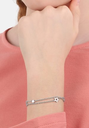 Bracciale argentato con due filamenti a catena, caratterizzato da charm a forma di stella e un gancio a moschettone. Indossato su un polso, contro una maglietta rosa.