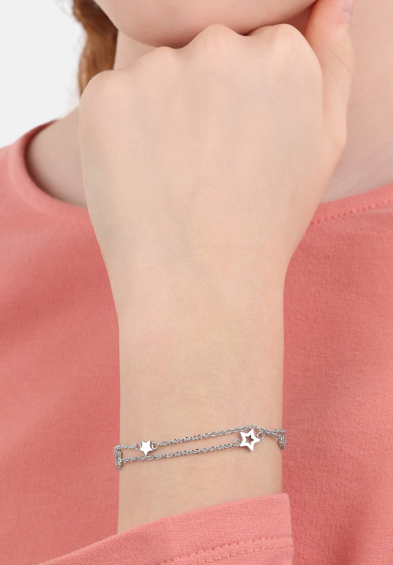 Zilveren armband met twee kettingstrengen, voorzien van stervormige bedels en een kreeftensluiting. Draagt om een pols, tegen een roze top.