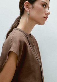 Bruine linnen blouse met een knoopsluiting en korte mouwen, gecombineerd met een zilveren hangoorbel met een sferisch detail.