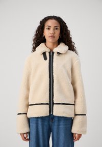 Cream-colored fleece jacket with a zip front, black accents, a collar, and wide sleeves. Features visible stitching and a boxy shape.