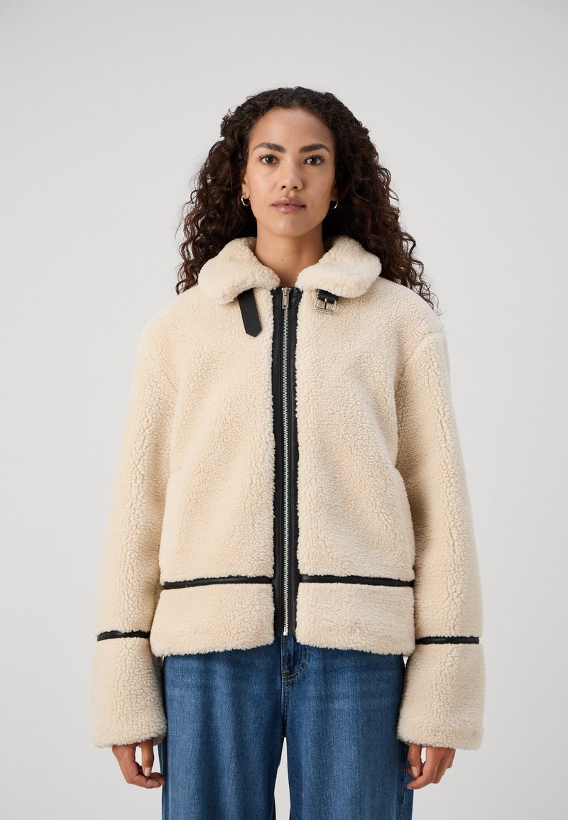 Cream-colored fleece jacket with a zip front, black accents, a collar, and wide sleeves. Features visible stitching and a boxy shape.