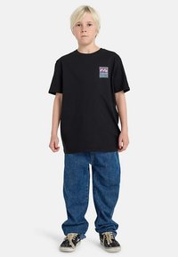 Musta puuvillainen t-paita, jossa on lyhyet hihat ja pieni monivärinen kuvio rinnassa. Yhdistetty löysälahkeisiin sinisiin farkkuihin ja mustiin lenkkareihin.