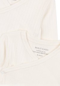 Chemise pour bébé en coton bio de couleur crème, texture côtelée, avec un col en V et une fermeture à boutons-pression. L'étiquette de taille indique 6 mois.