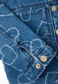 Denimjack met een lichtblauwe wassing, voorzien van gestikte hartpatronen en gouden knopen. Gedetailleerde naadconstructie en textuur zichtbaar.