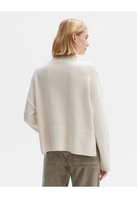 Cremefarbener Pullover mit hohem Kragen, geripptem Saum und überschnittenen Schultern. Weiche Textur, lockere Passform und leicht verkürzter Schnitt hinten.