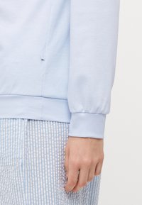 Ljusblå långärmad topp med en mjuk textur, elastiska manschetter och en liten sidospalt, tillsammans med blå- och vitrandiga byxor.
