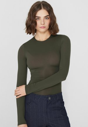 Kobieta o krótkich brązowych włosach, ubrana w dopasowaną oliwkowozieloną bluzkę z długim rękawem i ciemnoniebieskie spodnie w prążki, pozująca z założonymi na siebie ramionami.