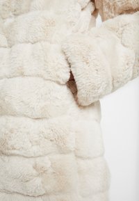 Faux fur-kappa i krämfärg, med kviltat mönster, plyschig textur och sidofickdetalj. Mjuk, tät tyg ger värme.