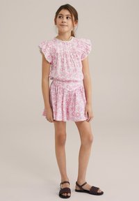 Rozā ziedu kleita ar frillēm un elastīgu vidukli; teksturēta auduma; kopā ar tumši brūnām pinumu sandālēm.