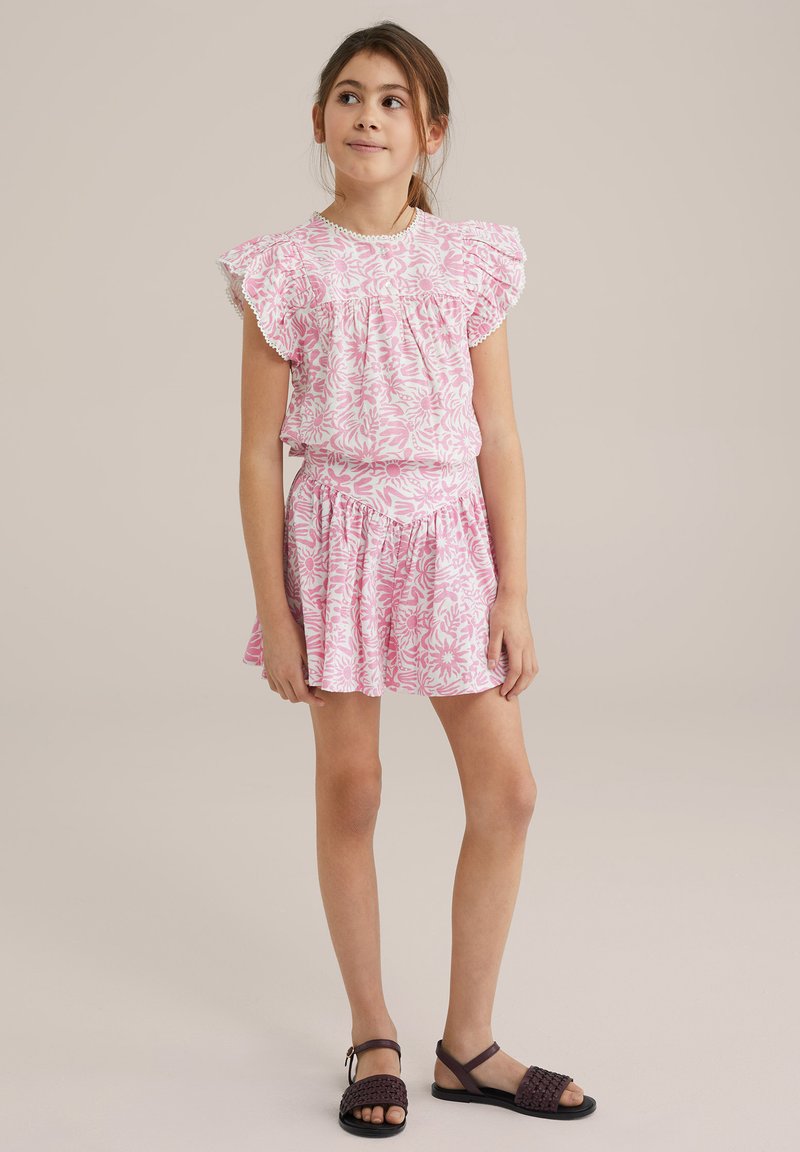 Rozā ziedu kleita ar frillēm un elastīgu vidukli; teksturēta auduma; kopā ar tumši brūnām pinumu sandālēm.