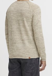 Man wearing beige textured long-sleeve sweater and faded black jeans, shown from the back against a plain white background.