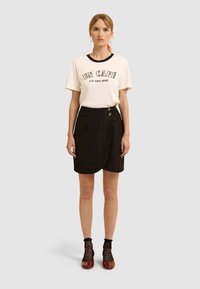 Kobieta ubrana w kremowy t-shirt z grafiką z napisem "UN CAFÉ s'il vous plaît", czarną spódnicę z zawinięciem, pończochy siatkowe do kostek oraz czerwone lakierowane buty typu Mary Jane.