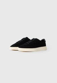 Par med lavtoppede svarte semskede sneakers med svarte lisser og off-white gummisåler på hvit bakgrunn.