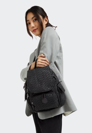 Black backpack with a textured pattern, multiple pockets, top flap, and a round logo detail. Worn on the shoulder against a light gray blazer.