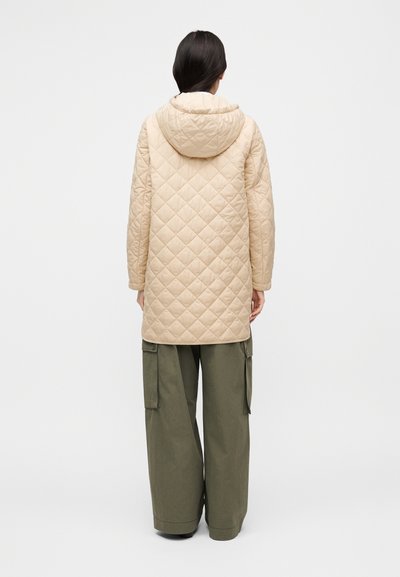 Manteau long matelassé beige avec capuche, présentant un motif en losanges, associé à un pantalon cargo olive ample. Fond uni.