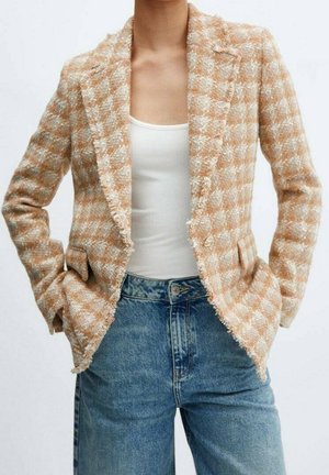 Personne portant un blazer en laine à carreaux beige et blanc par-dessus un débardeur blanc et un jean bleu, les mains dans les poches du blazer.