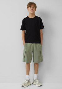 Schwarzes Baumwoll-T-Shirt kombiniert mit hellgrünen Cargoshorts, die über Seitentaschen verfügen, ergänzt durch weiße Socken und klobige graue Sneakers.