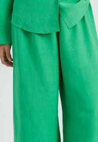 Groene linnen broek met een losse pasvorm, zichtbare textuur en subtiele glans. De tailleband is elastisch, wat comfort en bewegingsvrijheid biedt.