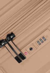 Valise rigide beige dotée de doubles fermetures éclair, d'un cadenas à combinaison TSA et d'une surface texturée pour plus de durabilité.