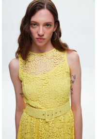 Gelbes Spitzenkleid mit Blumenmustern, ärmellosem Design und einem Gürtel mit quadratischer Schnalle. Glatte Textur mit einem leichten, luftigen Look.