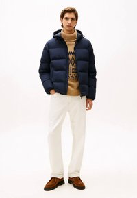 Marineblå dunjakke med hette, lys brun mønstret genser under, hvite bukser og brune sko. Uformelt antrekk med et varmt design.