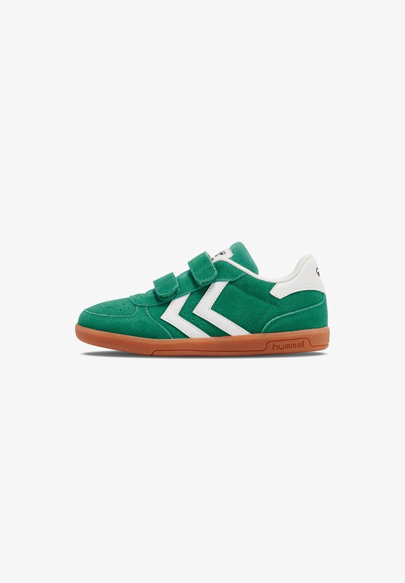La sneaker en daim vert présente des accents en chevron blancs, des doubles sangles en Velcro et une semelle en caoutchouc marron. Le logo est visible à l'arrière.