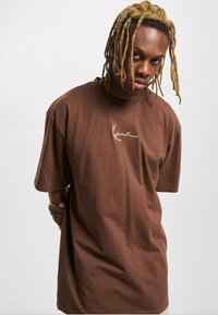 Camiseta de algodón marrón con un corte relajado, mangas cortas y un logotipo blanco escrito a mano en el pecho. Textura suave, diseño minimalista.