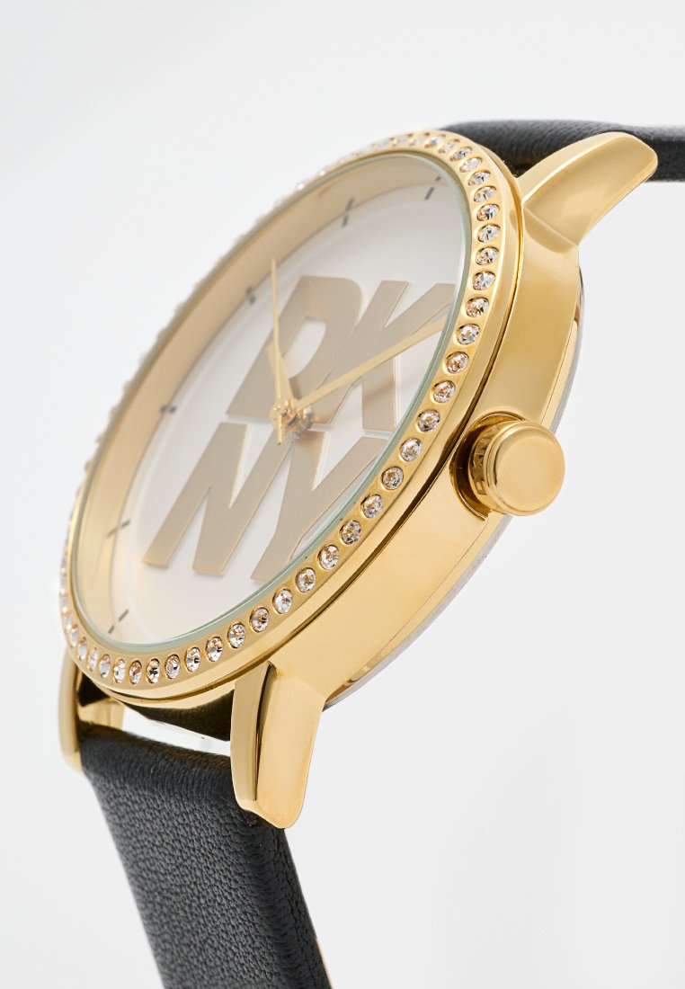 Goldfarbene Uhr mit großem weißem Ziffernblatt, das die Buchstaben "DKNY" zeigt. Schwarzes Lederarmband, Lünette verziert mit klaren Kristallen und einer Krone.