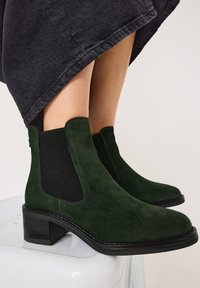 Dunkelgrüne Wildlederstiefeletten mit schwarzen elastischen seitlichen Einsätzen und einem niedrigen Blockabsatz, die eine glatte Textur und ein elegantes Design zeigen.