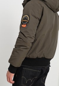 Olivenfarvet dunjakke med hætte, ribbet sort kant og en rund Superdry-logo på venstre skulder. Glat tekstur.