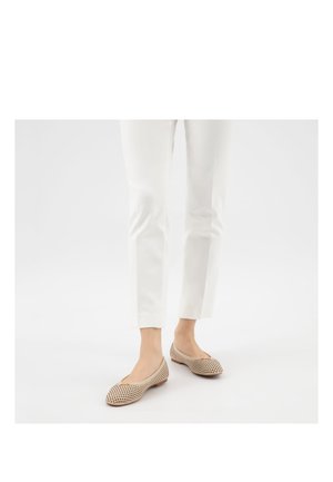 Person trägt weiße Hose und beige perforierte Ballerinas mit kleinem Schleifchen, steht vor weißem Hintergrund.