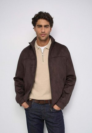 Hombre con cabello rizado que lleva una chaqueta marrón oscuro sobre un jersey beige con cremallera y vaqueros oscuros, con las manos en los bolsillos contra un fondo liso.