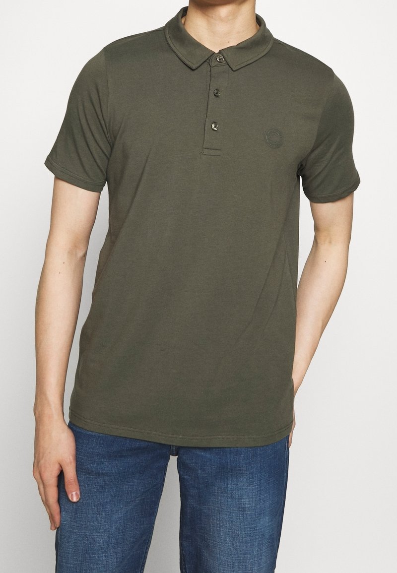 Olivově zelená polo tričko z bavlny, s klasickým límcem, třemi knoflíky na zapínání a jemným logem na hrudi. Nosené s džíny.