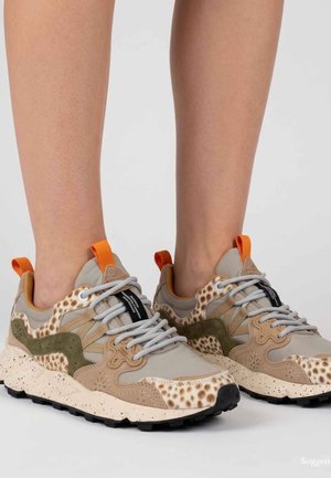 Sneakers beige e marroni con stampa animalier, lacci grigi e linguette arancioni indossate a piedi nudi su uno sfondo chiaro e uniforme.