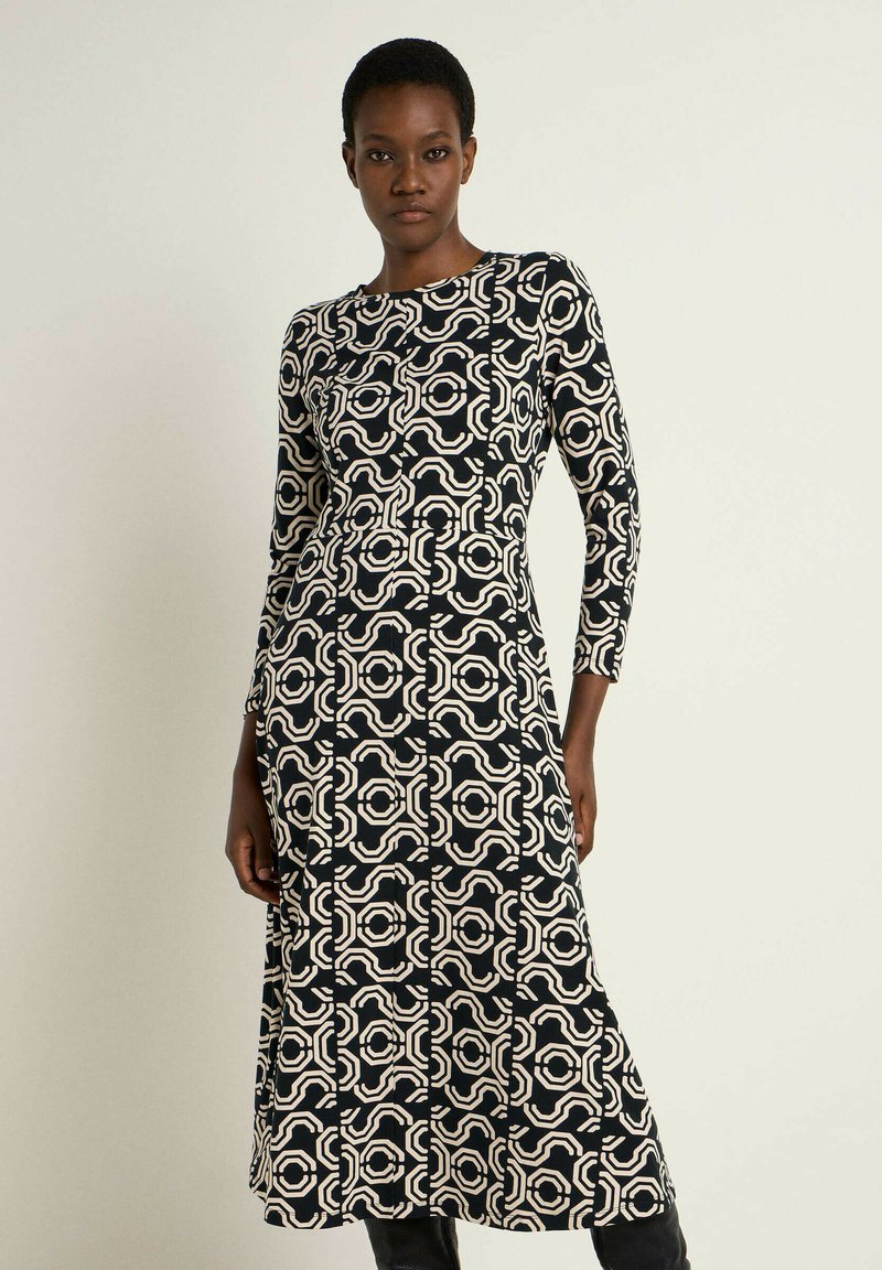 Schwarzes und cremefarbenes gemustertes Kleid mit langen Ärmeln, rundem Ausschnitt und tailliertem Oberteil, das ein geometrisches Design aufweist.