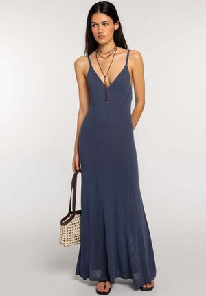 Mujer con un vestido maxi azul marino sin mangas con cuello en V, sosteniendo un bolso tejido y llevando collares en capas y sandalias.