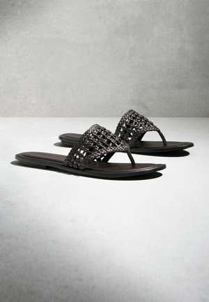 Čierne ploché sandále s otvorenou špičkou, s širokými, tkanými remienkami a palcovými prstencami na svetlosivej ploche a pozadí.