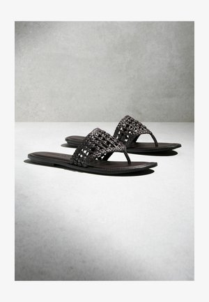 Čierne ploché sandále s otvorenou špičkou, s širokými, tkanými remienkami a palcovými prstencami na svetlosivej ploche a pozadí.