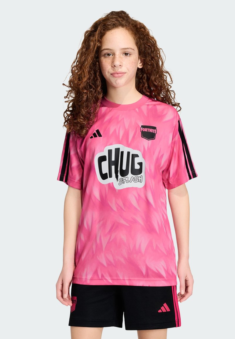 Jeune personne aux cheveux roux bouclés portant un maillot rose Fortnite Chug Splash et un short noir avec des rayures roses, debout devant un fond uni.
