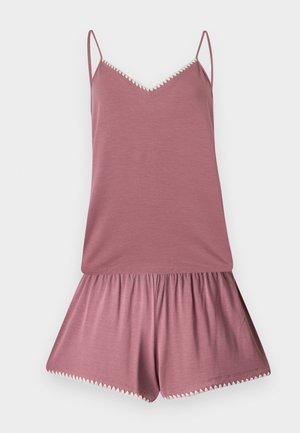 Ung kvinde står barfodet iført en støvet lilla camisole og matchende shorts med hvide kanter på en ensartet lys baggrund.