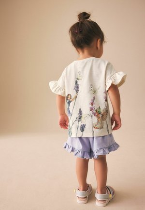 Kleinkind mit Dutt, trägt ein weißes Shirt mit floralem Feen-Print, lavendelfarbene Rüschenshorts und weiße Sandalen, steht auf beigem Boden.