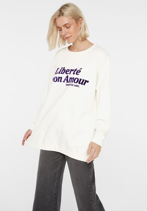 Blonde Frau trägt übergroßen weißen Pullover mit violettem Text "Liberté mon Amour" und lockeren dunkelgrauen Jeans, steht vor weißem Hintergrund.
