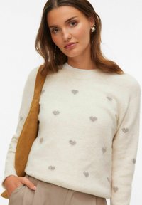 Pull couleur crème avec un col rond et des motifs de cœurs argentés. Le matériau est doux, avec une coupe décontractée. Porté avec un sac à bandoulière marron.