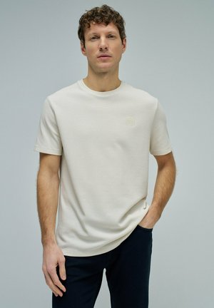 Hombre con cabello rizado que lleva una camiseta texturizada de manga corta color crema y pantalones oscuros, de pie con una mano en el bolsillo contra un fondo liso.