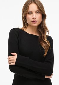 Zwarte top met lange mouwen, zachte textuur en brede halslijn, uitgevoerd in een aansluitend ontwerp. Achtergrond is effen wit.