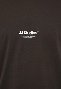 Καφέ βαμβακερό t-shirt με λευκό λογότυπο "JJ Studios" και το κείμενο "ΠΑΡΟΥΣΙΑΖΕΤΑΙ ΑΠΟ ΤΗΝ ΟΜΑΔΑ J&J™ ΣΧΕΔΙΑΣΜΕΝΟ ΣΤΗΝ ΔΑΝΙΑ."