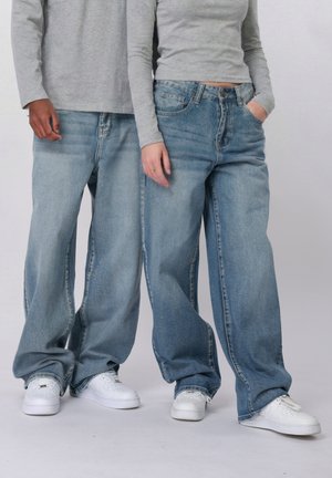 Kaks inimest, kes kannavad helehallis pikka varrukaga toppe, lahtisi helerohelisi teksapükse ja valgeid tenniseid, seistes kõrvuti ühevärvilise tausta ees.