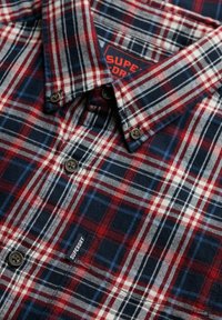 Camisa de cuadros rústica hecha de franela, con colores rojo, marino y blanco. Incluye cierres de botones y una pequeña etiqueta de marca en la parte delantera.
