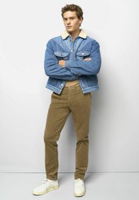 Jeansjacke mit Lammfellkragen, kombiniert mit olivgrünen Cordhosen und weißen Sneakern. Das Modell steht mit verschränkten Armen vor einem schlichten Hintergrund.