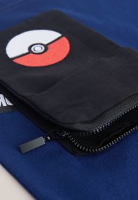 Petite pochette noire zippée avec un motif Poké Ball rouge et blanc reposant sur un fond de tissu bleu plié.